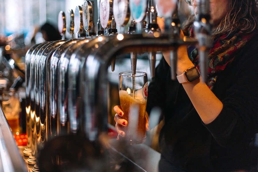 A woman pulls a pint from one of several beer taps on the cover image for Legal Drinking Age In London.