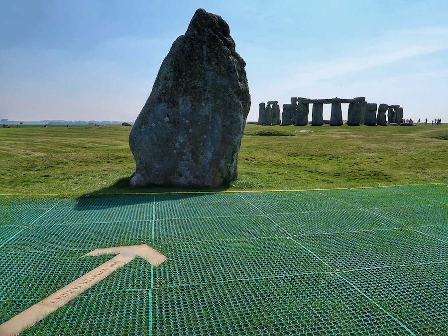 A rock with an arrow on the ground pointing towards Stonehenge in the background.