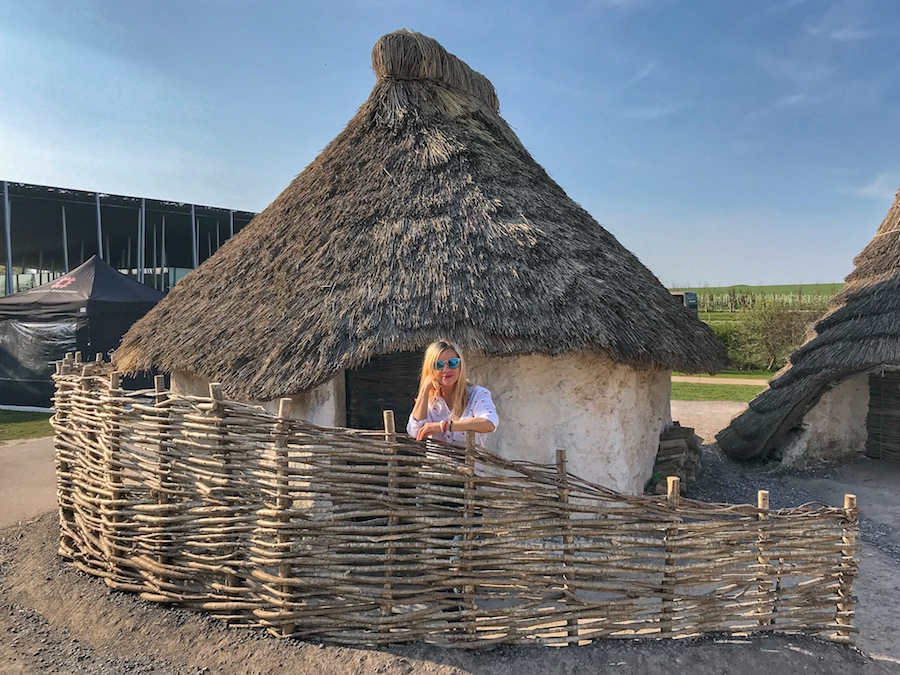 Zuzi is standing in front of a Neolithic house at Stonehenge. The house is white, small and has a straw roof