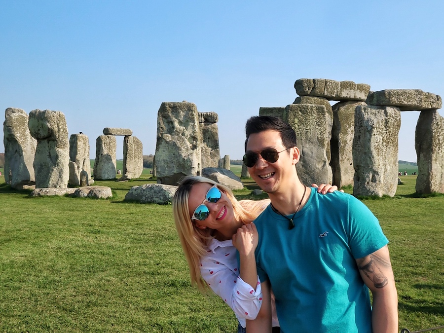 Stonehenge stone circle on a clear day, with two visitors smiling in the foreground and the standing stones visible behind them on Salisbury Plain.